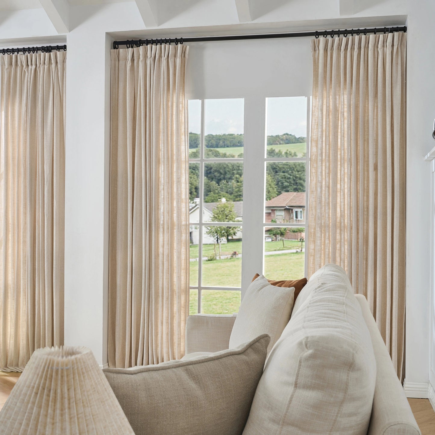 Light beige poly viscose linen window drapes, soft curtains and a sofa in a room with a large window view.
