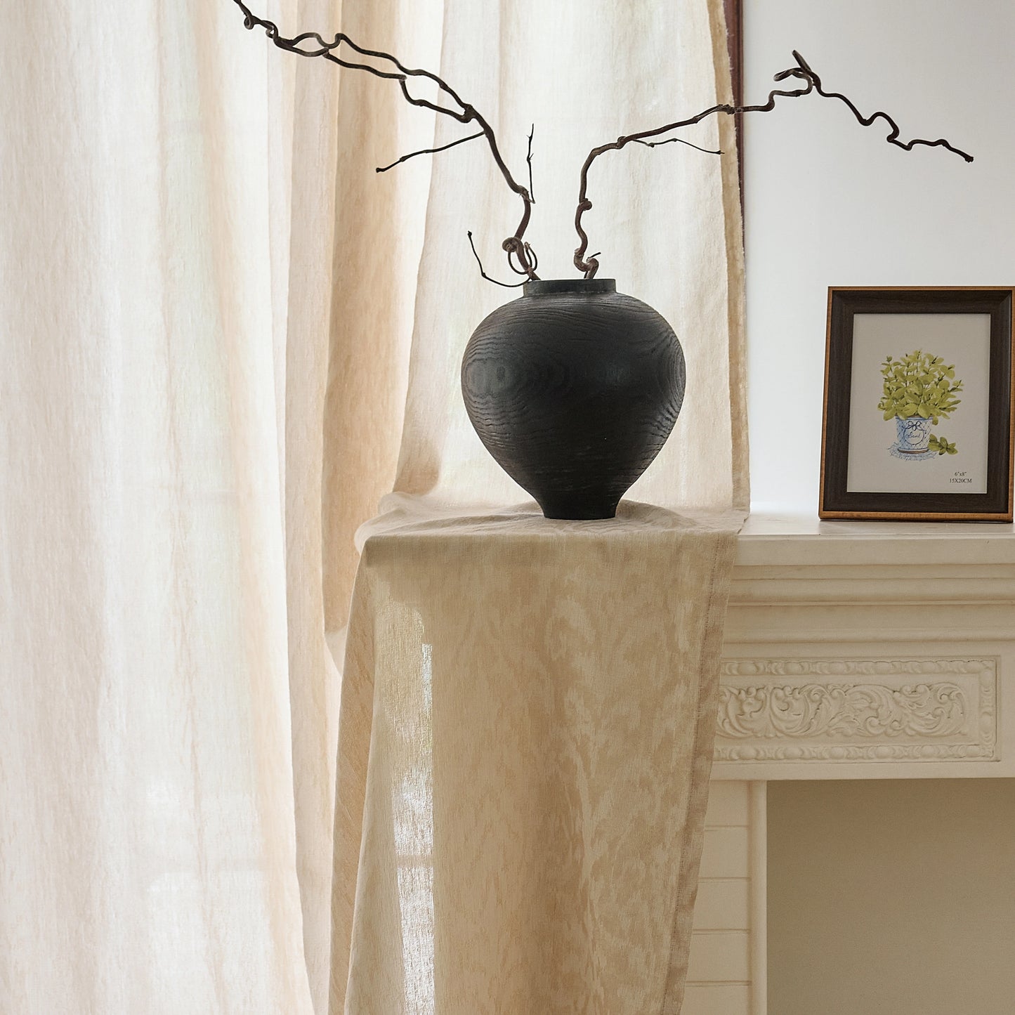 Black vase on table with light-beige wool gauze curtain featuring softly textured plant-inspired pattern in the background.