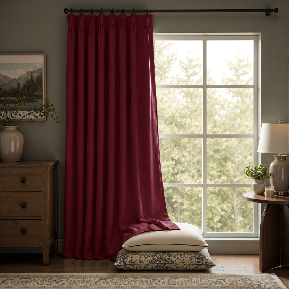 A deep burgundy curtain panel hanging on a black rod in a styled room with a window, wooden furniture, and pillows on the floor.