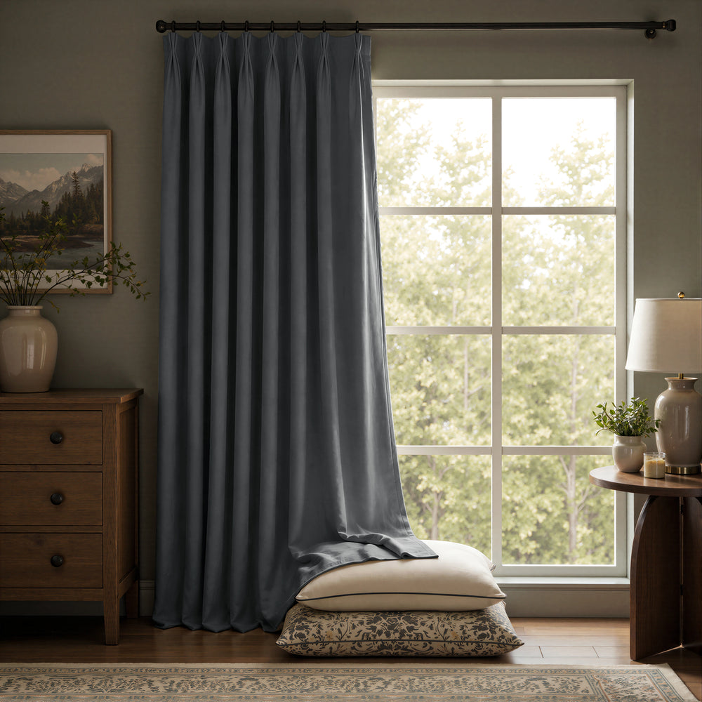 A long slate blue curtain panel hanging on a dark rod in a styled room with a window, wooden furniture, a lamp, and pillows on the floor.
