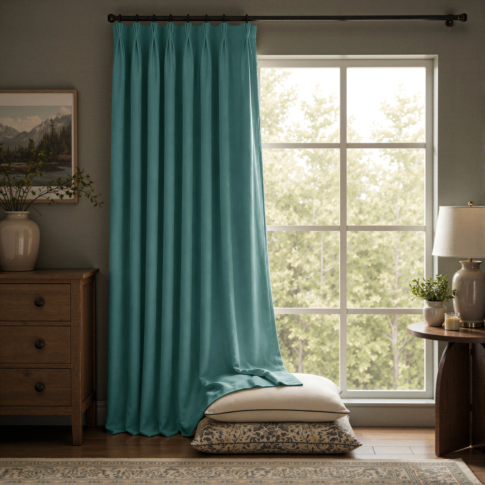 A single teal curtain panel hanging on a dark rod in a styled room with a large window, wood furniture, a lamp, and pillows on the floor.