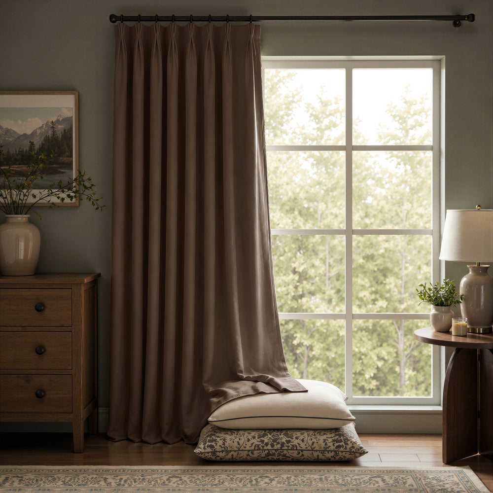 A pair of floor-length taupe brown curtains hanging on a black rod over a large multi-pane window, with pillows stacked on the floor in front. The room features a wooden dresser on the left and a side table with a lamp on the right.