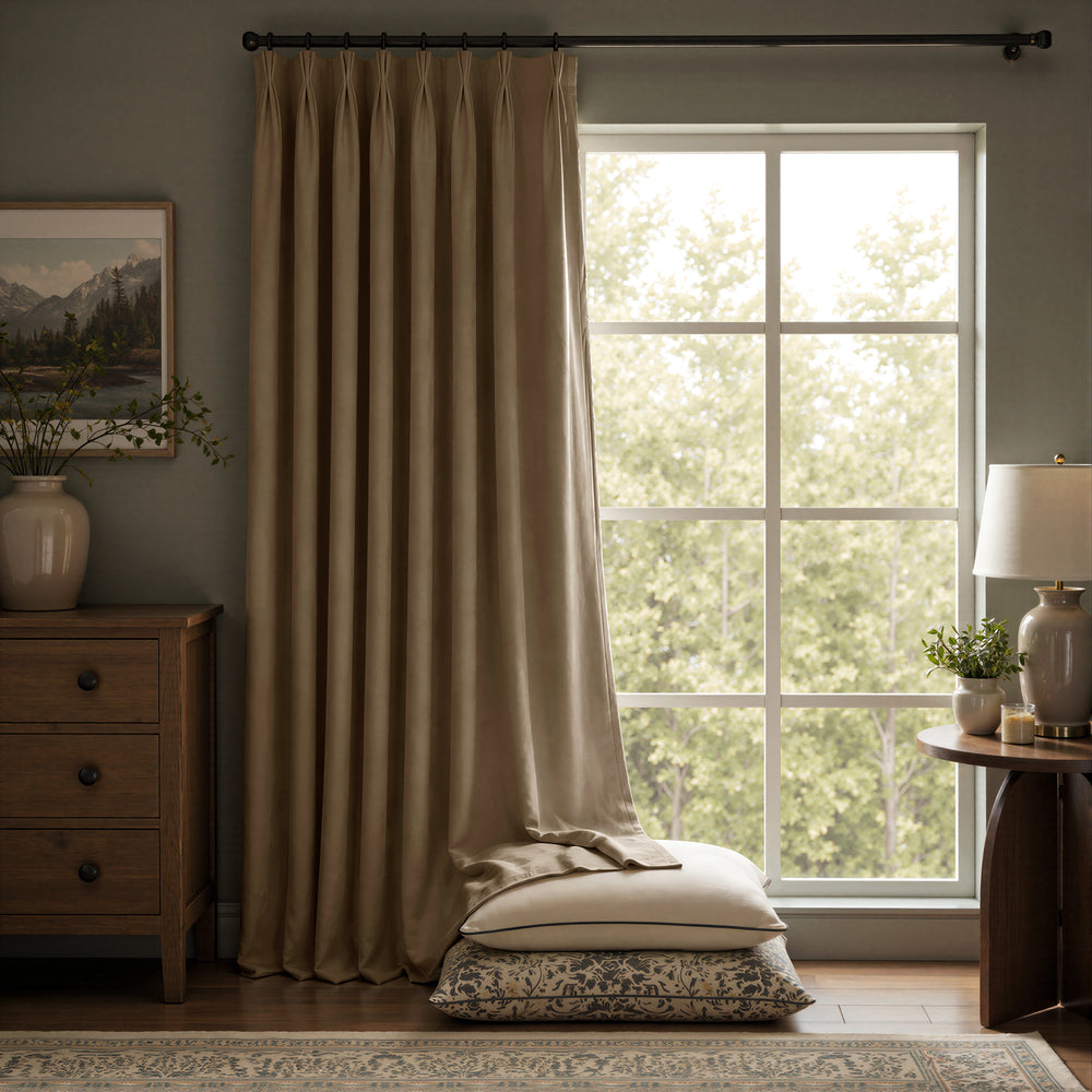 A pair of floor-length taupe curtains hanging on a black rod over a large multi-pane window, with a matching folded pillow and patterned cushion stacked on the floor in a styled room setting.