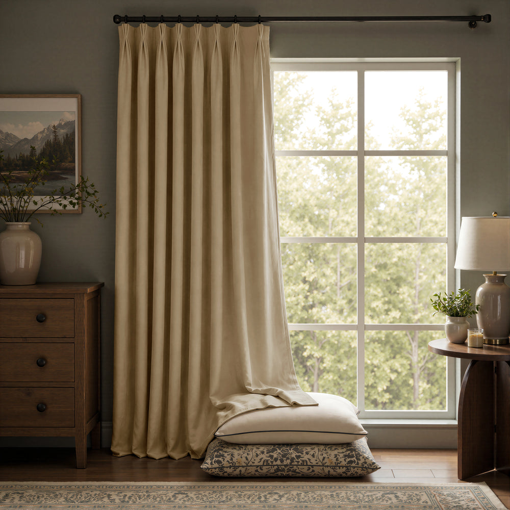 A pair of floor-length beige curtains hanging on a black rod over a large multi-pane window, with pillows stacked on the floor in front. The room has a muted blue-gray wall, a wooden dresser on the left, and a side table with a lamp on the right.