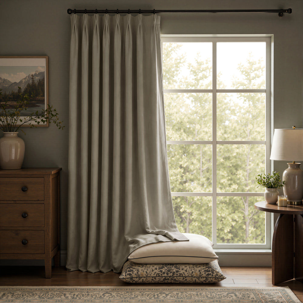 A pair of light sage green curtain panels hanging on a black rod in a styled room with a window, wooden furniture, and a lamp.