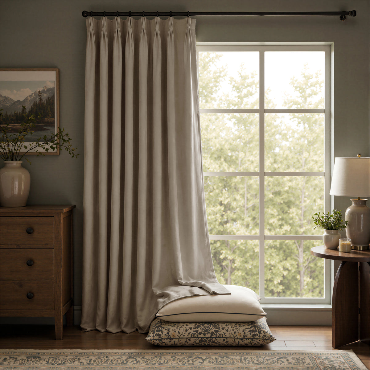 A pair of long taupe-beige curtain panels hanging on a black rod in a styled room with a window, wooden furniture, and a lamp.
