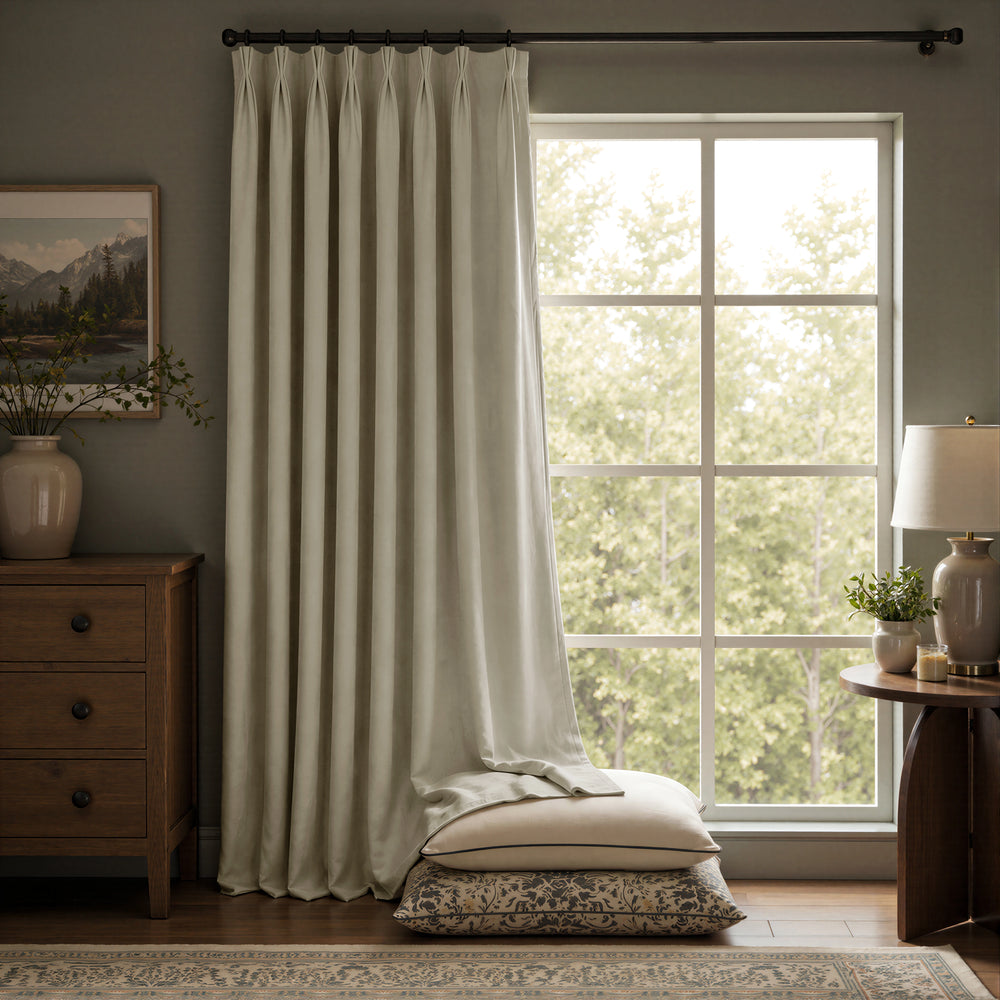 A pair of floor-length curtain panels in a light warm gray or off-white tone hanging on a dark rod in a styled room with a window, wooden furniture, and pillows on the floor.