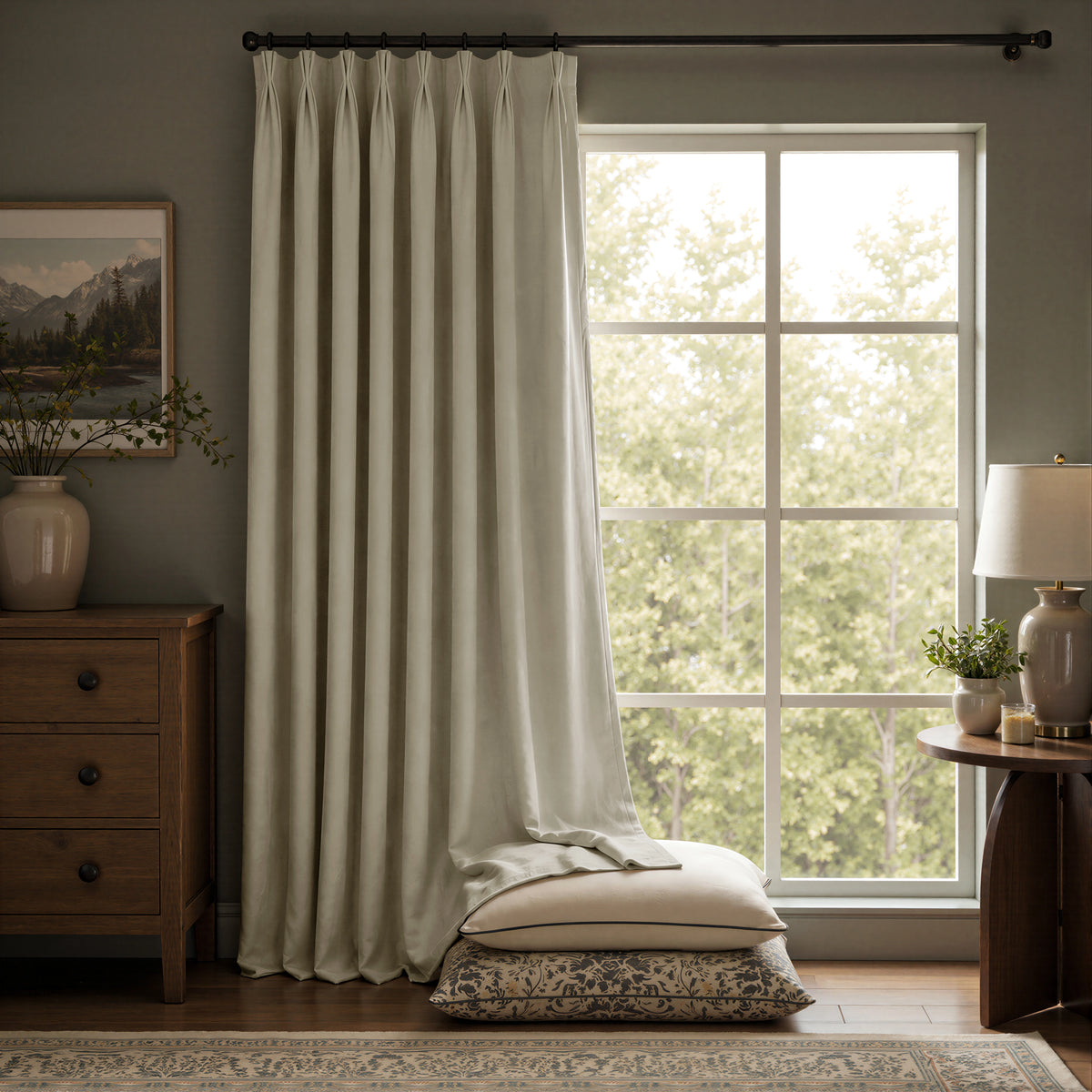A pair of floor-length curtain panels in a light warm gray or off-white tone hanging on a dark rod in a styled room with a window, wooden furniture, and pillows on the floor.