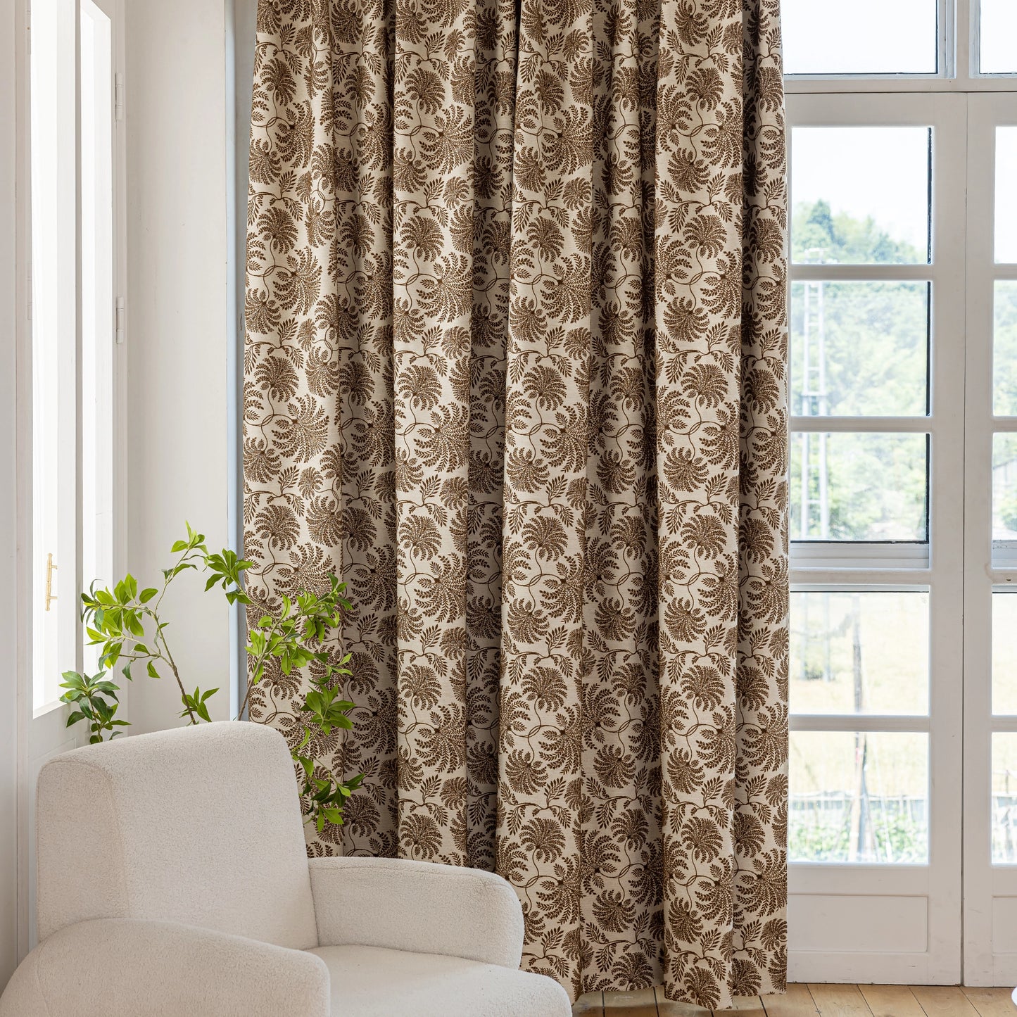 Decorative chocolate fern-inspired boho curtain in a room with a white armchair and windows.