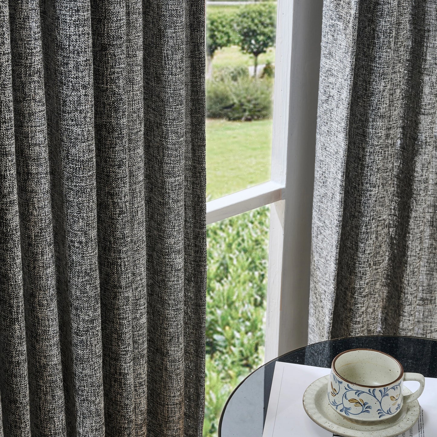 Dark gray and white textured poly viscose linen curtain with a cup and saucer on a table by a window overlooking greenery.