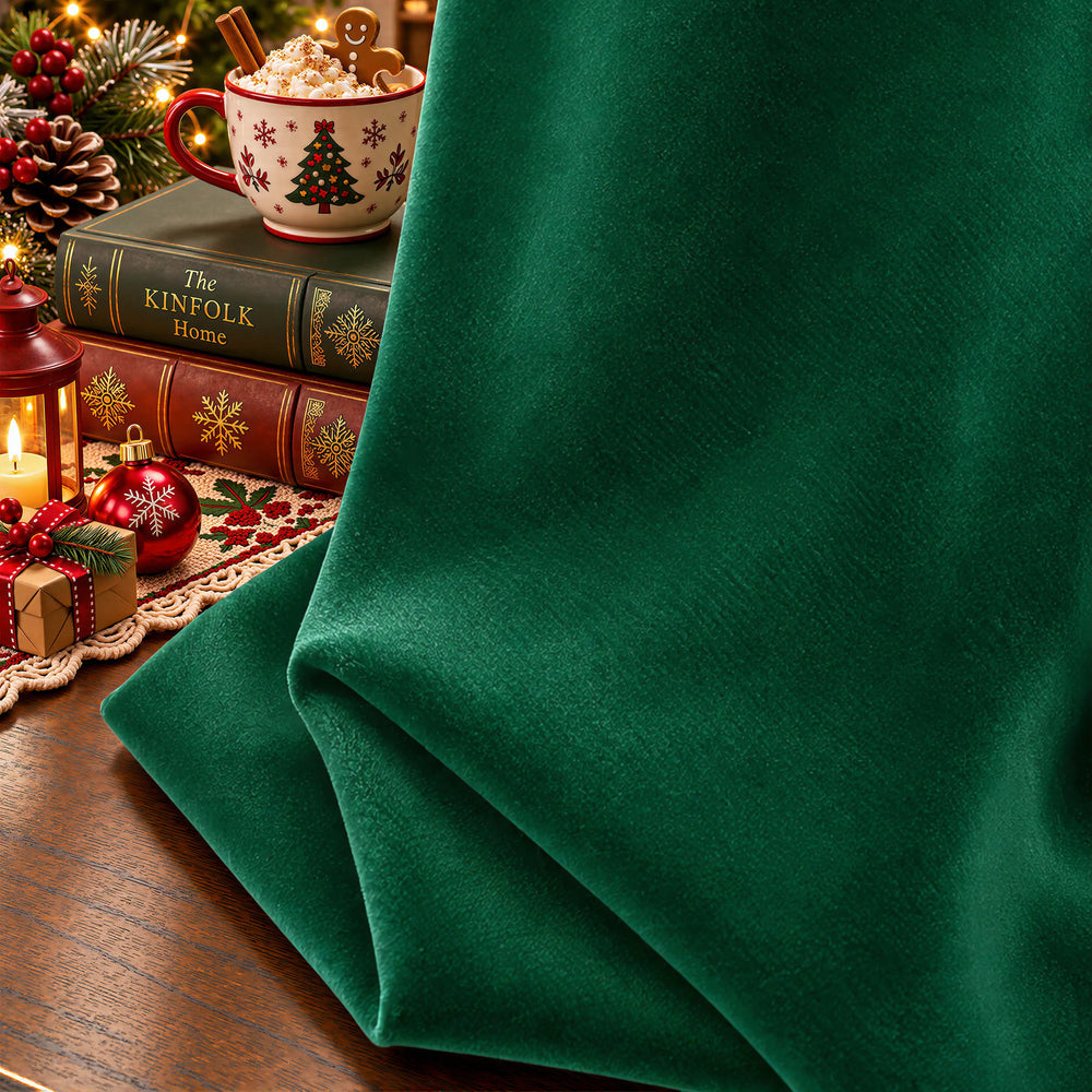 A close-up of rich emerald green velvet fabric draped over a wooden table, with a vintage teacup and stacked books in the softly blurred background.