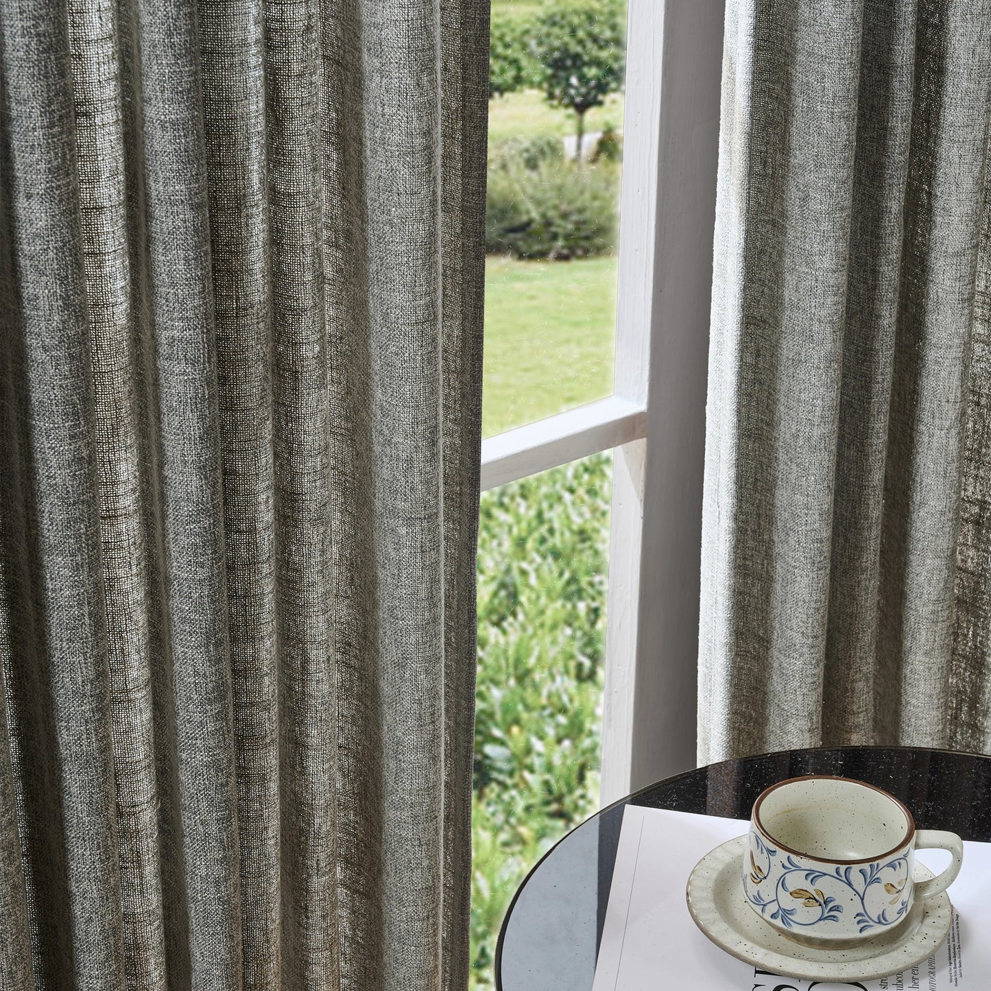 Heather gray textured poly viscose linen curtain with a cup and saucer on a table by a window overlooking greenery.
