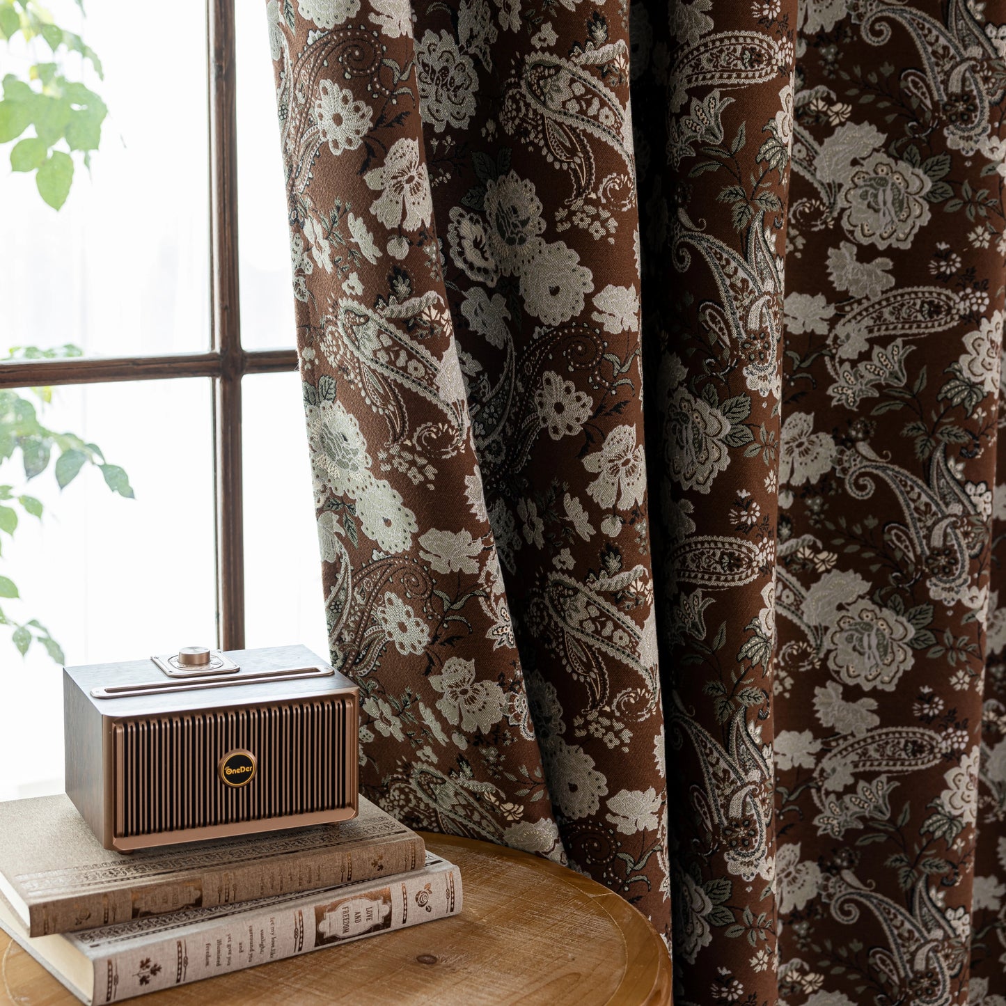 Decorative curtain with floral pattern in front of a window, with a small speaker on books in the foreground.