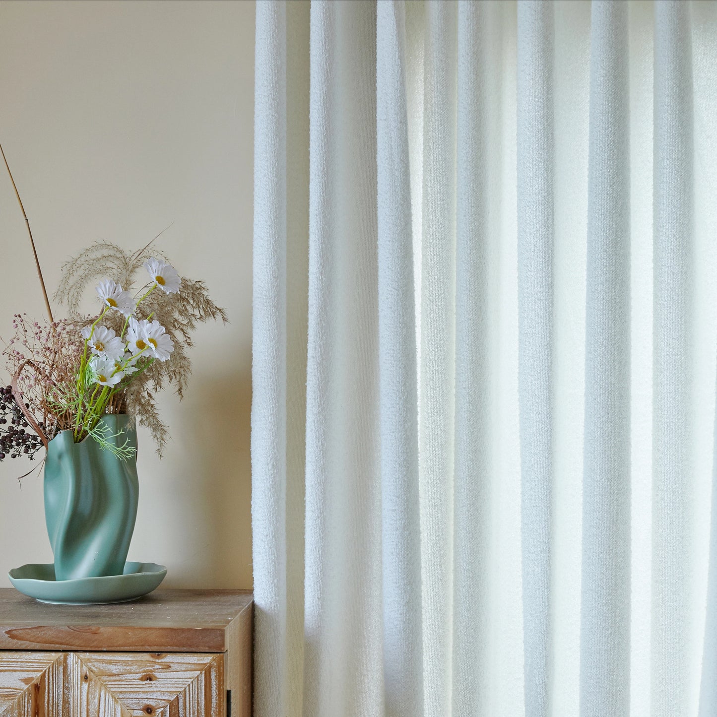 Teal vase with dried flowers on a wooden surface next to a close-up of off-white terry yarn curtain textile.