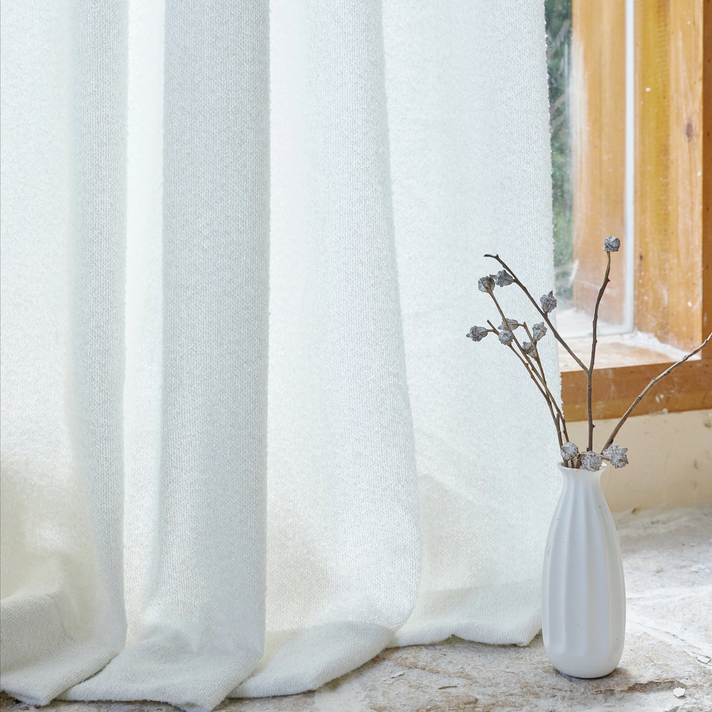 White terry yarn curtains with airy weave, with a white vase containing dried branches on the floor.