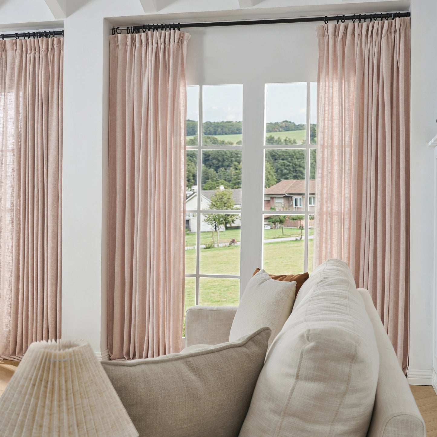 Pale pink poly viscose linen window drapes, soft curtains and a sofa in a room with a large window view.