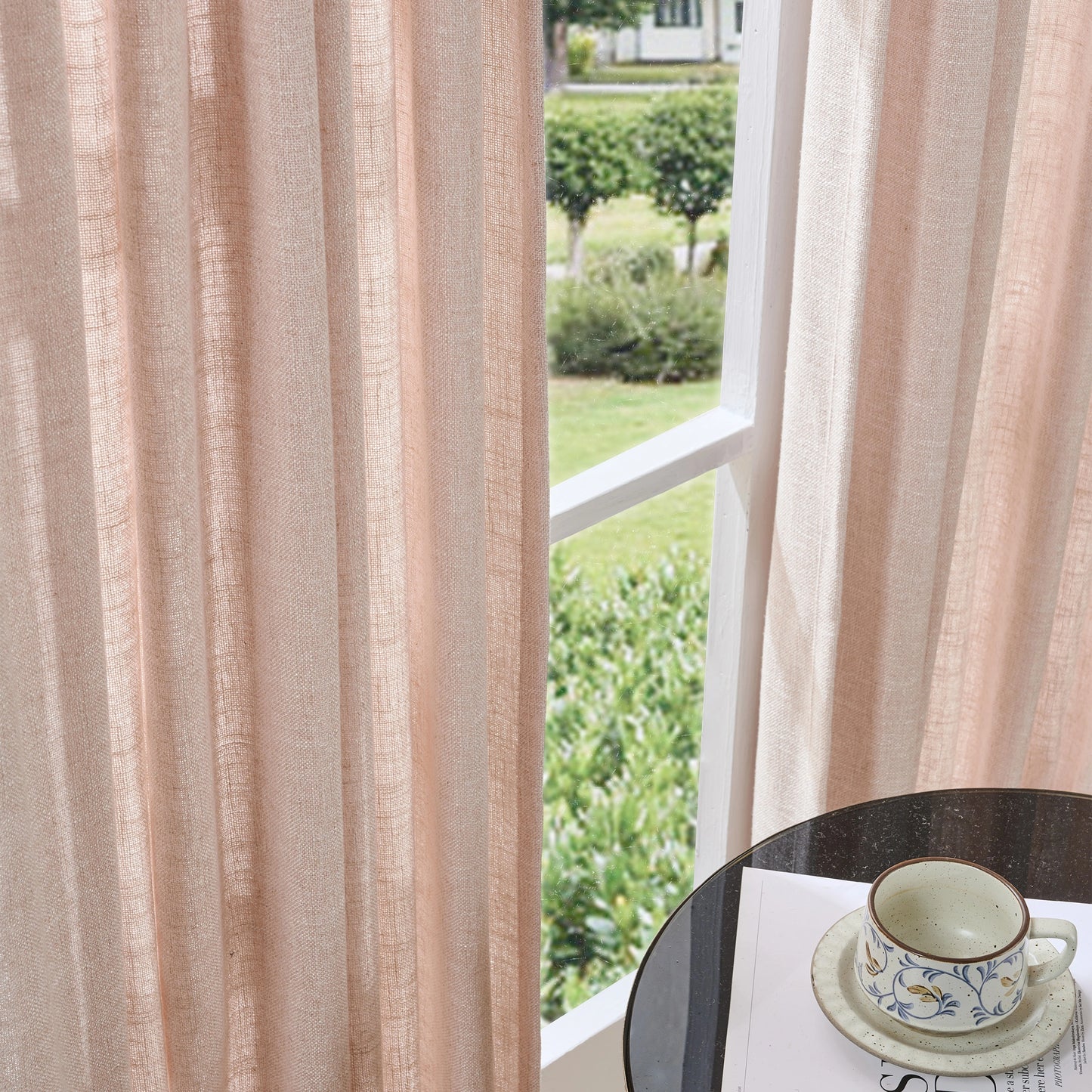 Pink-toned textured poly viscose linen curtain with a cup and saucer on a table by a window overlooking greenery.