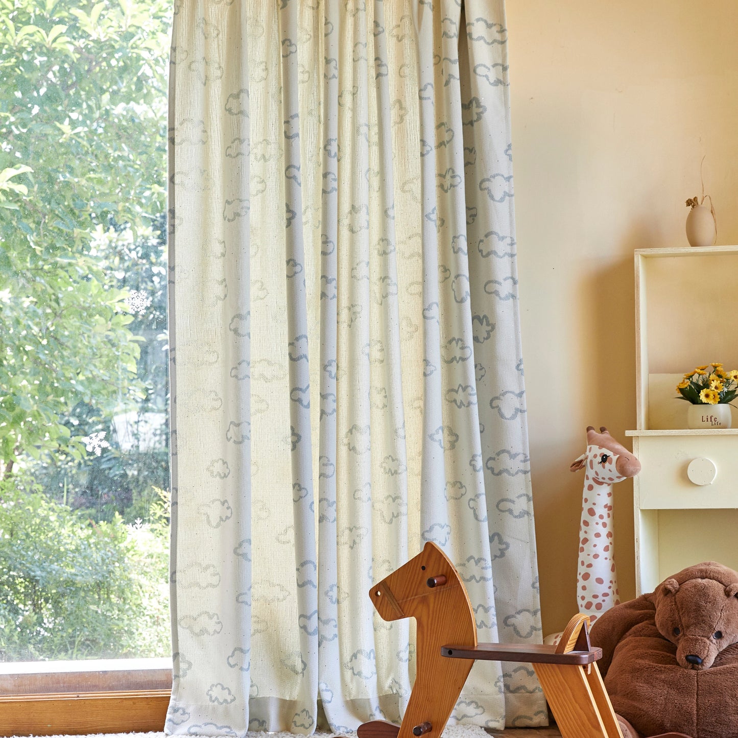 Children's room with a rocking horse, bear, and giraffe toys by a window with blue line white cloud drapes.