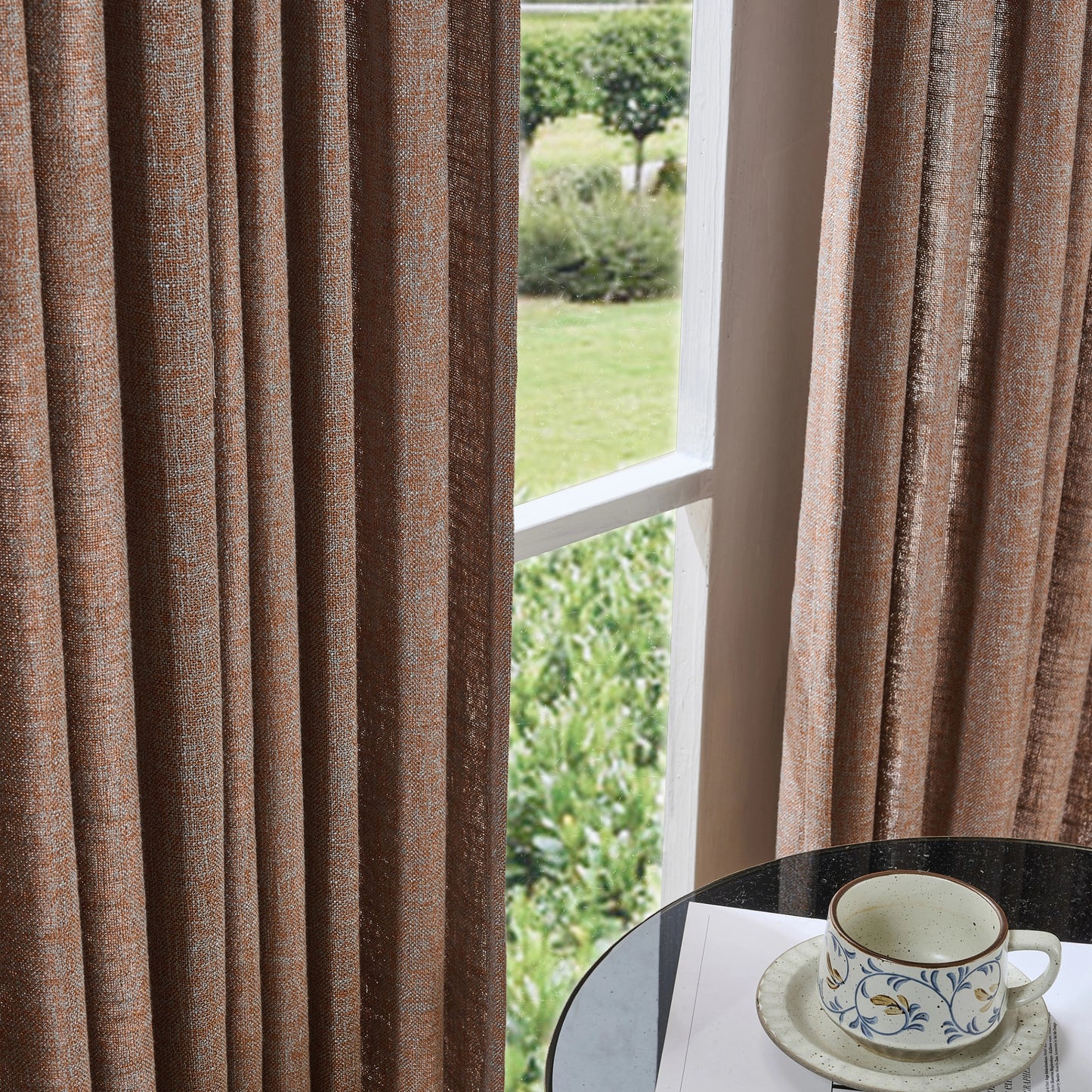 Muted rust brown textured poly viscose linen curtain with a cup and saucer on a table by a window overlooking greenery.