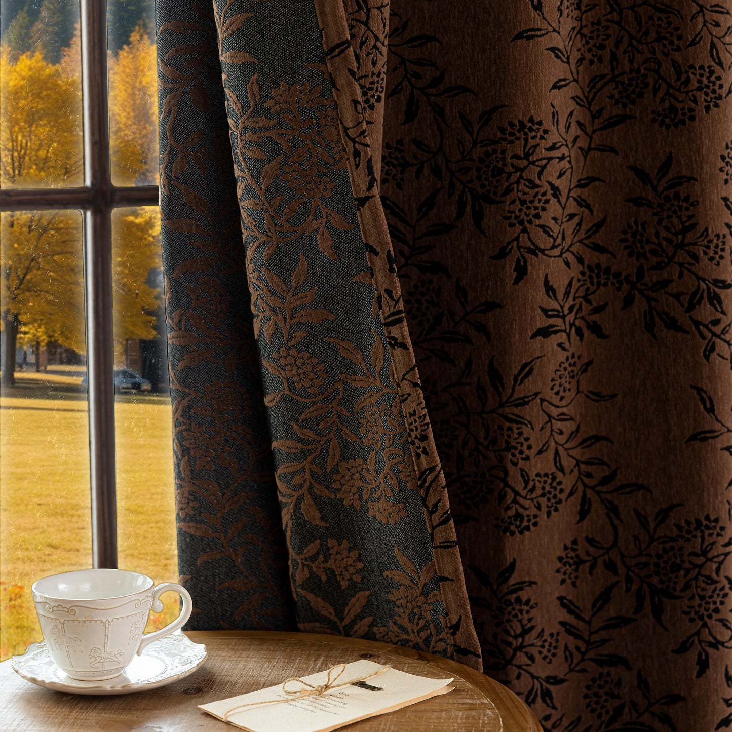Decorative curtains with a floral pattern in front of a window with autumn view, featuring a cup and saucer on a table.